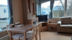 A cozy living room with a wooden dining table, chairs, a sofa, and a TV. There is a window with curtains, a ceiling fan, and a radiator. - Northfield 198, Skipsea Sands (Driffield)