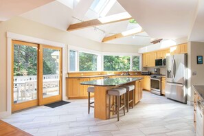 A modern kitchen with wooden cabinets, a central island with stools, a stainless steel refrigerator, and large windows. - Home by National Seashore with Bball Court! (Wellfleet)