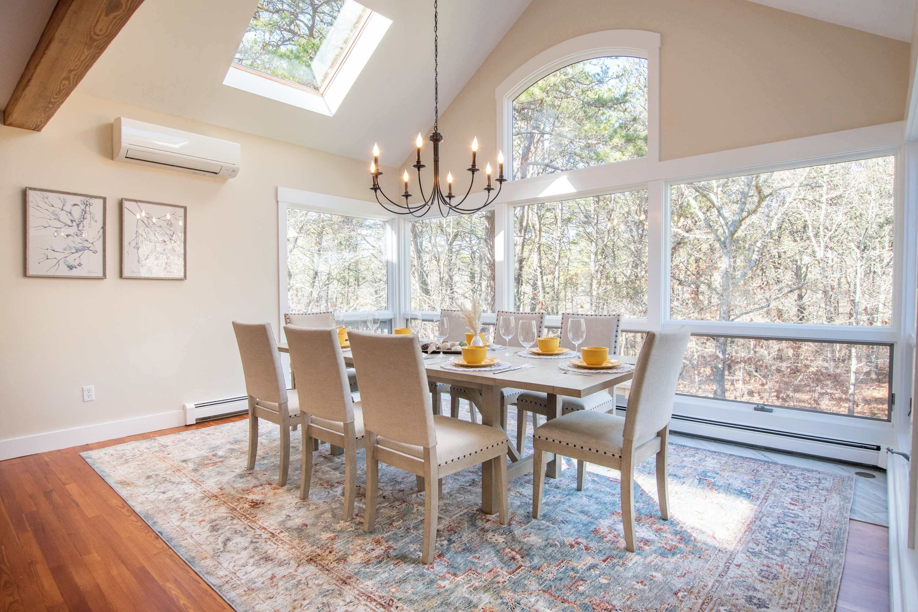 Une salle à manger avec une grande table, des chaises, un lustre et de grandes fenêtres donnant sur la verdure.