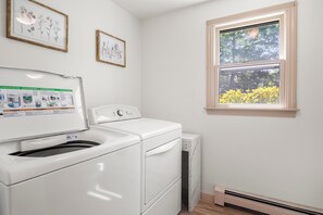 A laundry room with a front-loading washer and dryer, a window with a view of trees, and two framed botanical prints on the wall.