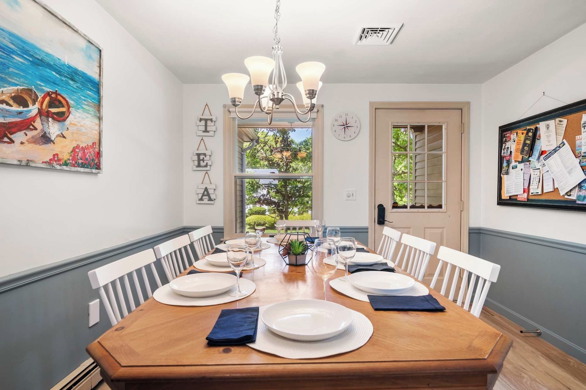 A dining area with a wooden table set for four, white chairs, a chandelier, and a painting of boats on the wall.