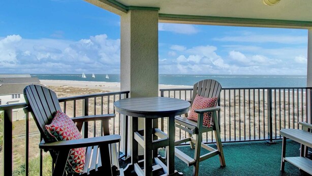 Varanda com duas cadeiras Adirondack e uma pequena mesa redonda, com vista para uma praia com veleiros e céu claro.