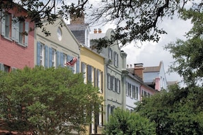A row of colorful buildings with shutters, a flag, and trees in the foreground. - 12 42nd Avenue by Avantstay 2 Mins To The Beach Screened In Porch! (Isle of Palms)
