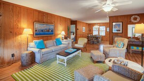 A cozy living room with wooden walls, a ceiling fan, a dining area, and a kitchen in the background. - 12 42nd Avenue by Avantstay 2 Mins To The Beach Screened In Porch! (Isle of Palms)