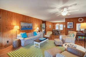 A cozy living room with wooden walls, a ceiling fan, a dining area, and a kitchen in the background. - 12 42nd Avenue by Avantstay 2 Mins To The Beach Screened In Porch! (Isle of Palms)