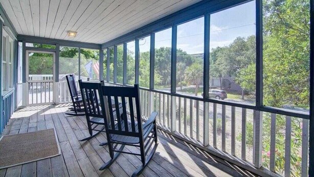 A covered porch with wooden flooring, black rocking chairs, and large windows offering a view of trees and a road. - 12 42nd Avenue by Avantstay 2 Mins To The Beach Screened In Porch! (Isle of Palms)