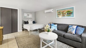 Uma sala de estar moderna com um sofá cinzento, uma mesa de centro branca e uma cama com cabeceira cinzenta.