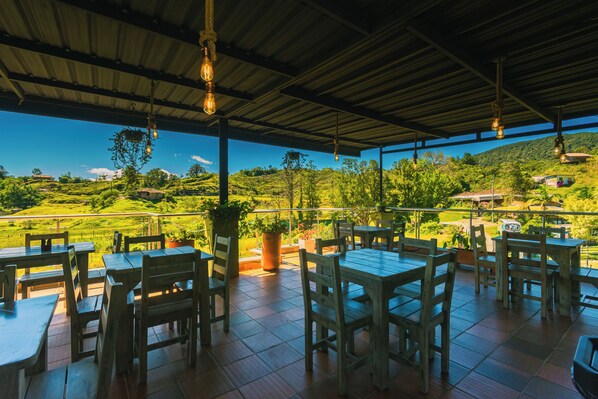 An outdoor patio with wooden tables and chairs, potted plants, and hanging light fixtures.