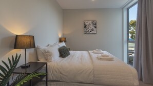 A neatly made bed with pillows and a fluffy pillow, a bedside table with a lamp, a wall-mounted picture, and a window with curtains.