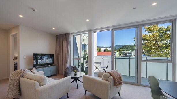 A modern living room with a sofa, armchair, and a small table. A television is mounted on the wall, and there is a balcony with a view of the city and hills.