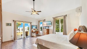 A spacious bedroom with a large bed, a ceiling fan, a television, and a view of the outdoors through a sliding door. - Villa el Encanto//Amazing views, walk to beach! (Rincon)