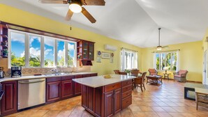 A spacious kitchen with a large island, wooden cabinets, and a dishwasher. There are large windows providing natural light and views of greenery outside. - Villa el Encanto//Amazing views, walk to beach! (Rincon)