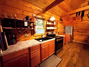 Cozinha de uma cabana de madeira, com micro-ondas, fogão, pia e prateleiras com diversos objetos.