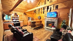 Ein gemütliches Blockhaus-Interieur mit Kamin aus Stein, Ledersitzmöbeln, einer Küchenzeile und einem an der Wand befestigten Fernseher.