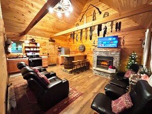 L’intérieur chaleureux d’un chalet avec une cheminée en pierre, des fauteuils en cuir, un coin cuisine et un téléviseur fixé au mur.