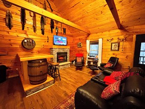 Interior de cabana de madeira com lareira de pedra, assentos de couro, televisão instalada na parede e um painel de peles de animais.