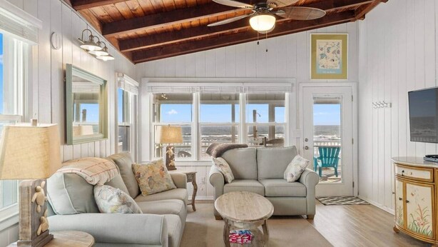 A cozy living room with a ceiling fan, large windows, and a view of the ocean. - Oak Island Beachfront Bungalow - Direct Ocean Views + Sleeps 8 (Oak Island)