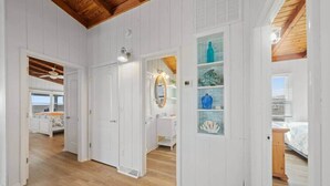 A hallway with wooden floors and white walls, leading to a bedroom with a bed and a bathroom with a sink and mirror. - Oak Island Beachfront Bungalow - Direct Ocean Views + Sleeps 8 (Oak Island)