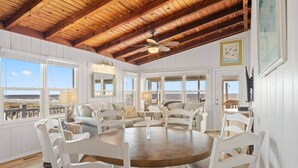 A dining area with a round table and white chairs, large windows with ocean views, and a wooden ceiling. - Oak Island Beachfront Bungalow - Direct Ocean Views + Sleeps 8 (Oak Island)