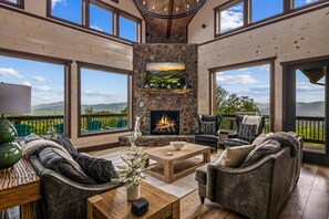 A spacious living room with a stone fireplace, large windows, and a view of the mountains.