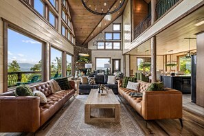 A spacious living area with leather sofas, a coffee table, and a fireplace. Large windows offer views of the outdoors.