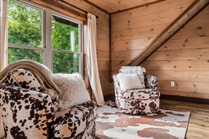 A cozy room with a wooden ceiling, a large window with curtains, a sofa with a fluffy pillow, and a chair with a patterned cushion.