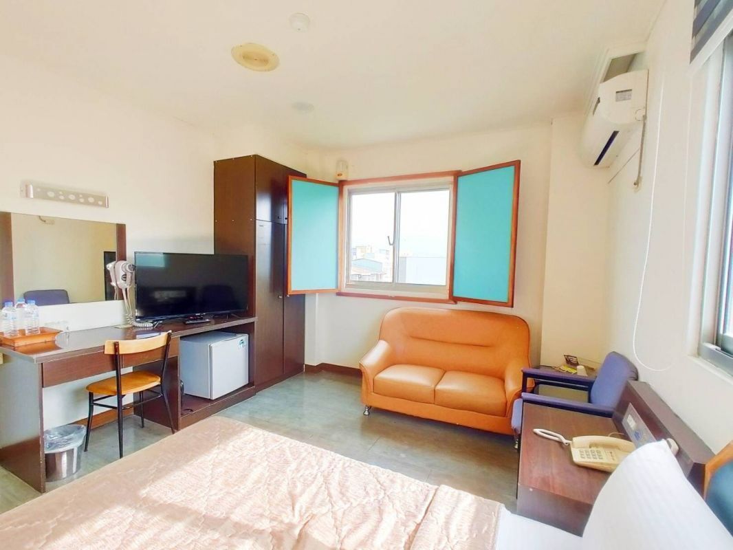 A hotel room with a bed, a desk with a television, a small refrigerator, an orange leather sofa, and a window with a view.
