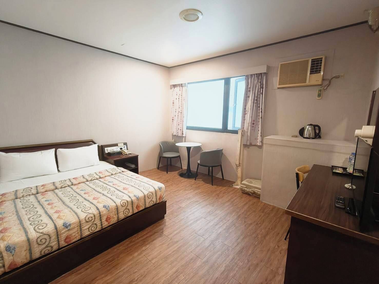 Una habitación de hotel con cama, escritorio, silla, mesita, radiador y ventana con cortinas.