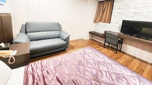 A hotel room with a grey sofa, a bed with a pink quilt, a TV on a wooden stand, and a stone-textured wall.