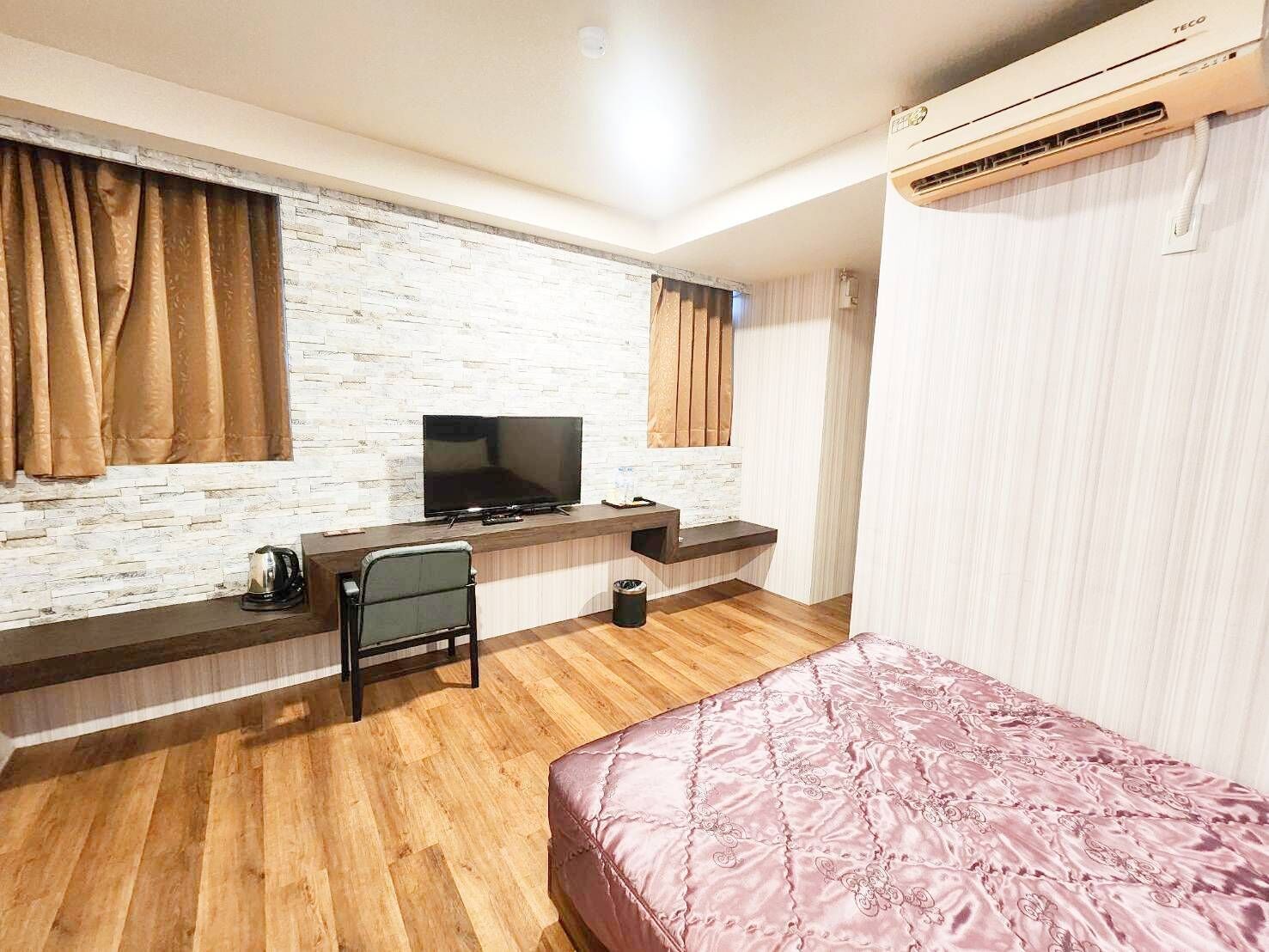A hotel room with a TV, a desk, a chair, a bed, and an air conditioning unit.
