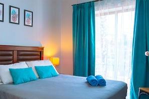 Quarto de hotel com uma cama, cortinas verde-água, mesa de cabeceira com abajur e obras de arte emolduradas na parede.