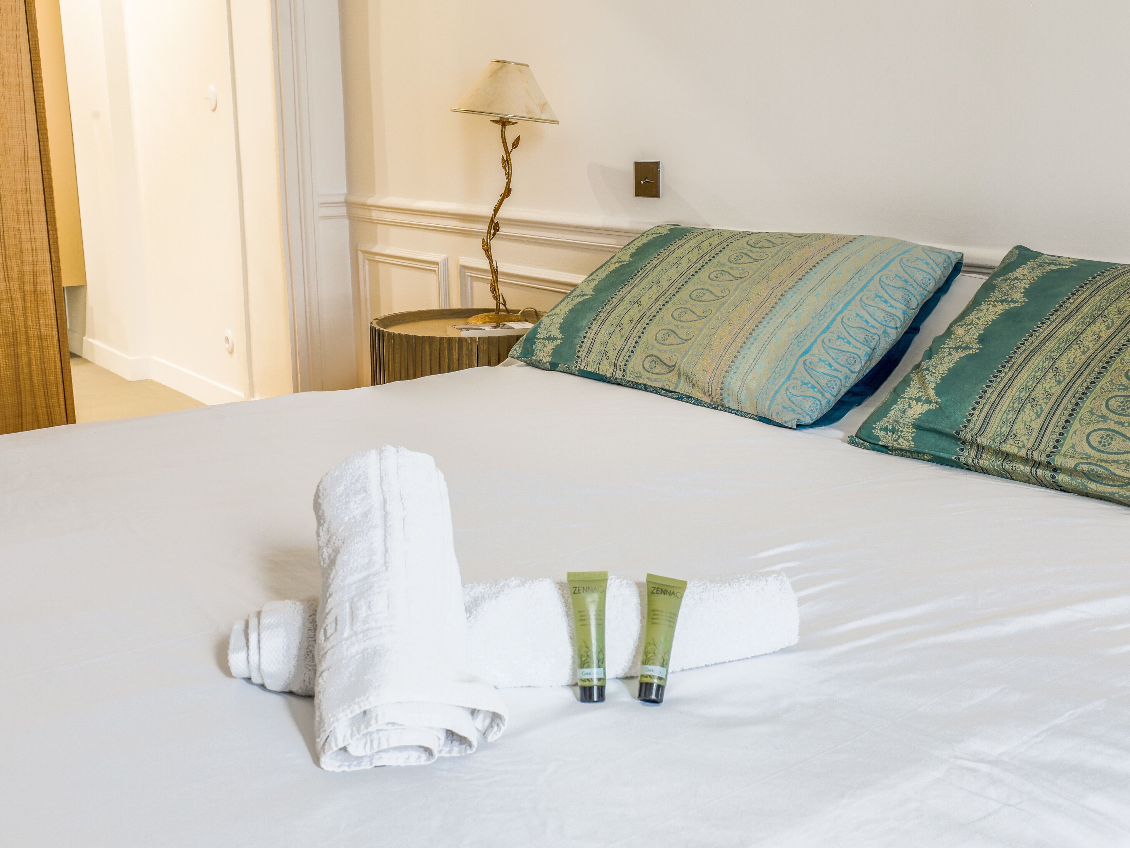 Una cama bien hecha con una toalla blanca enrollada, dos tubos de loción, un cojín estampado de color verde y una lámpara en una mesita de noche.