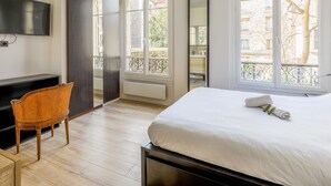 A hotel room with a bed, a TV, a chair, and large windows.