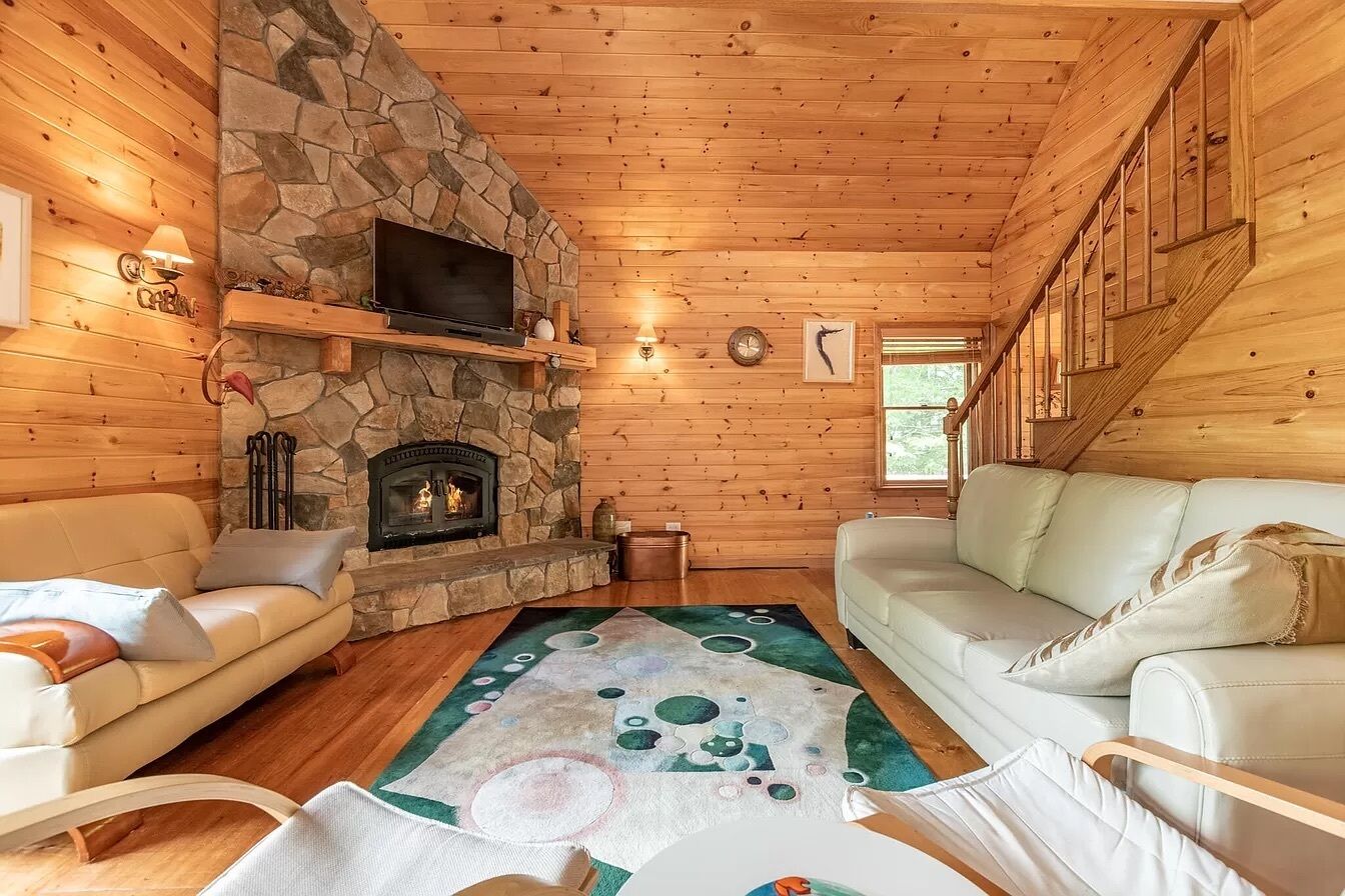 A cozy cabin interior with a stone fireplace, wooden walls, a flat-screen TV, and two sofas.
