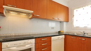 A kitchen with wooden cabinets, a dishwasher, an oven, and a window with a floral curtain. - CANIGO: House with private pool in l'Escala ideal for families (L'Escala)