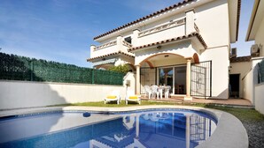A swimming pool with a clear blue water surface, surrounded by a white pool deck. In the background, there is a two-story building with a tiled roof and a balcony. - CANIGO: House with private pool in l'Escala ideal for families (L'Escala)
