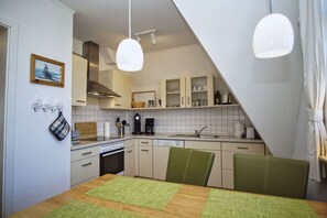 A compact kitchen with a dining area, featuring a wooden table, green chairs, a coffee maker, and a clock on the wall. - Hafenhäuser Breege Wohnung Nr. 03 Blinkfeuer - Hafenhäuser Breege (Breege)