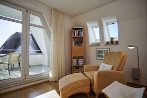 Un salon chaleureux avec une grande fenêtre, une bibliothèque, un fauteuil confortable et un lampadaire.