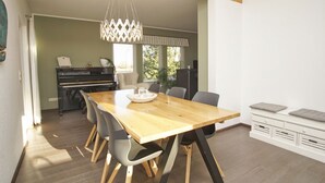 A dining area with a wooden table, grey chairs, a chandelier, and a piano in the background. - Haus Sternschnuppe - comfortable house with fireplace, sauna, garden, terrace - (Wiek)