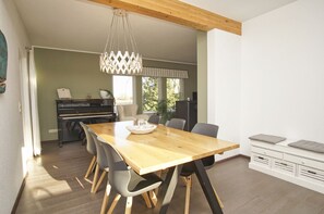 A dining area with a wooden table, grey chairs, a chandelier, and a piano in the background.