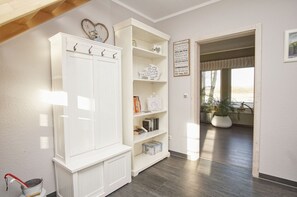 A spacious room with a white wardrobe, open shelving with books and decorative items, a view through an open doorway to a balcony with plants, and a sign on the wall.