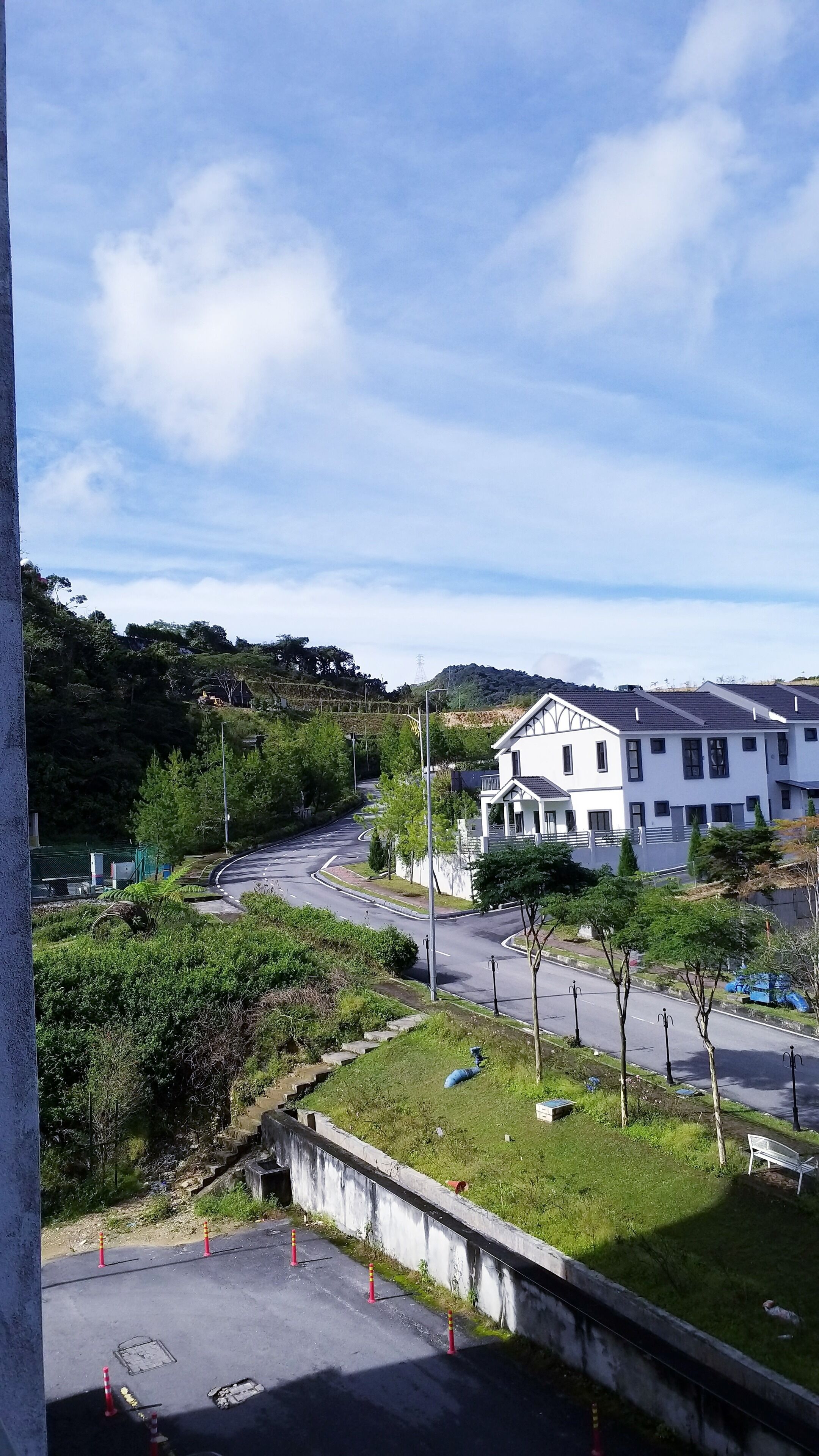A residential area with a white two-story house, a curved road, and a grassy hill in the background.