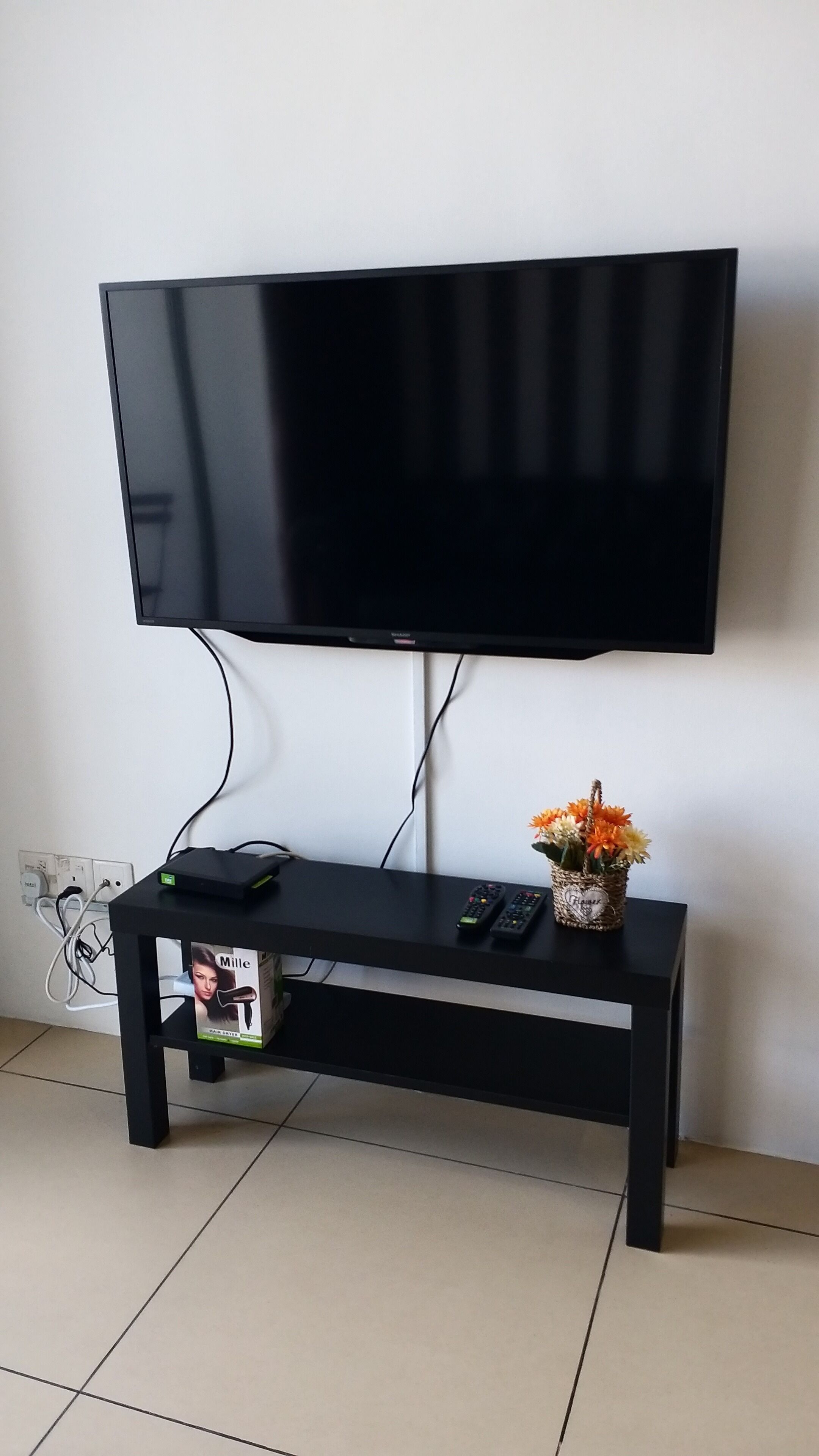 Un mueble bajo negro para televisor con un televisor de pantalla plana montado encima. Hay un control remoto y un pequeño florero con flores sobre el mueble.