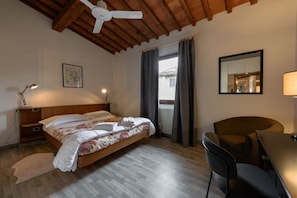 Une chambre avec un plafond en bois, un lit recouvert dâune couvre-lit Ă motifs floraux, un bureau avec une chaise, un lampadaire et une fenĂȘtre avec des rideaux.