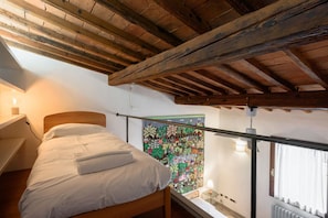 A bedroom with a wooden ceiling, a bed with white bedding, a colorful tapestry on the wall, and a window with curtains.