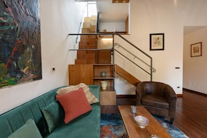 Un salon avec un canapé vert, un fauteuil en cuir marron, une table basse en bois, un escalier avec une rampe en métal et un tableau abstrait aux couleurs vives accroché au mur.