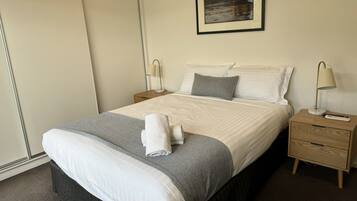 A neatly made bed with a grey and white duvet, two bedside lamps, and a small wooden nightstand with a remote control.