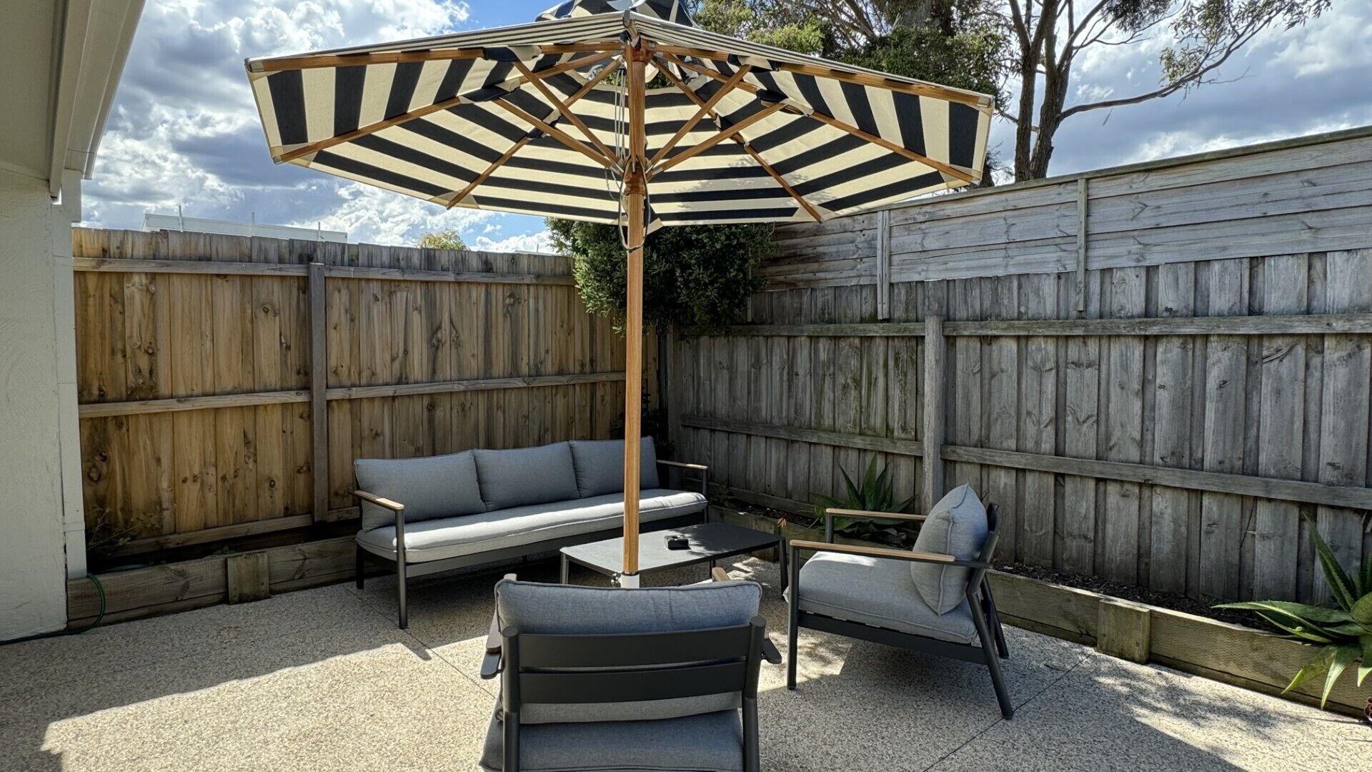 Un patio con cerco de madera, un sofá gris, una silla y un paraguas a rayas.