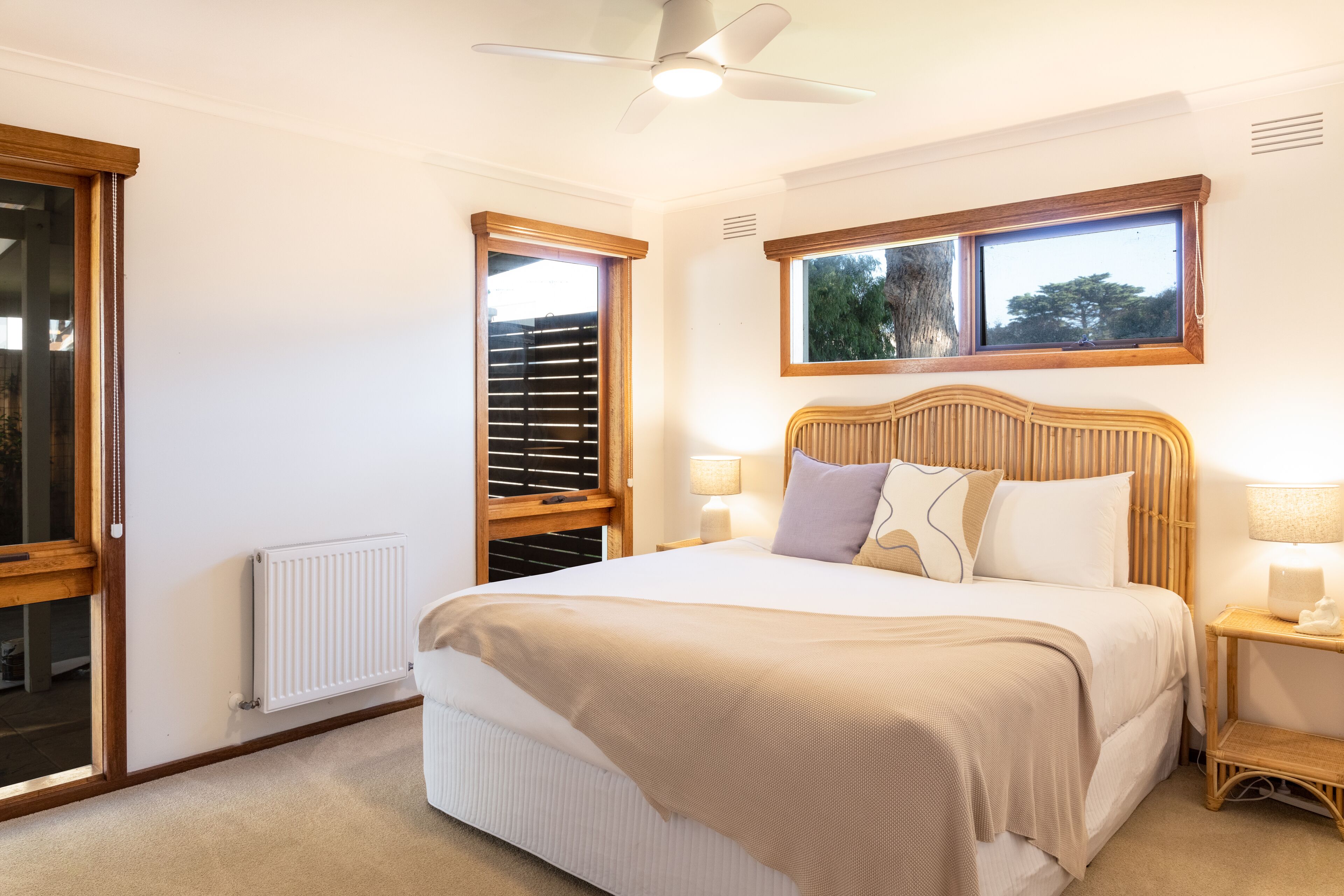 Un dormitorio con una cama grande, cabecera de madera, dos lámparas de noche, una ventana con persianas y un ventilador de techo.
