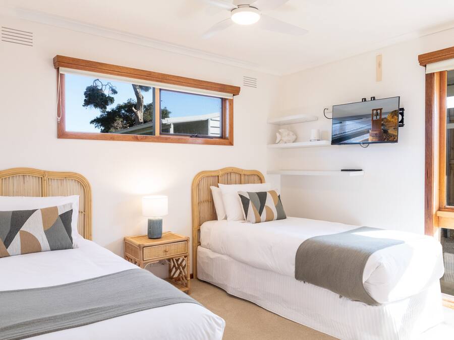 A room with two beds, a ceiling fan, a TV mounted on the wall, and a window with a view of trees.